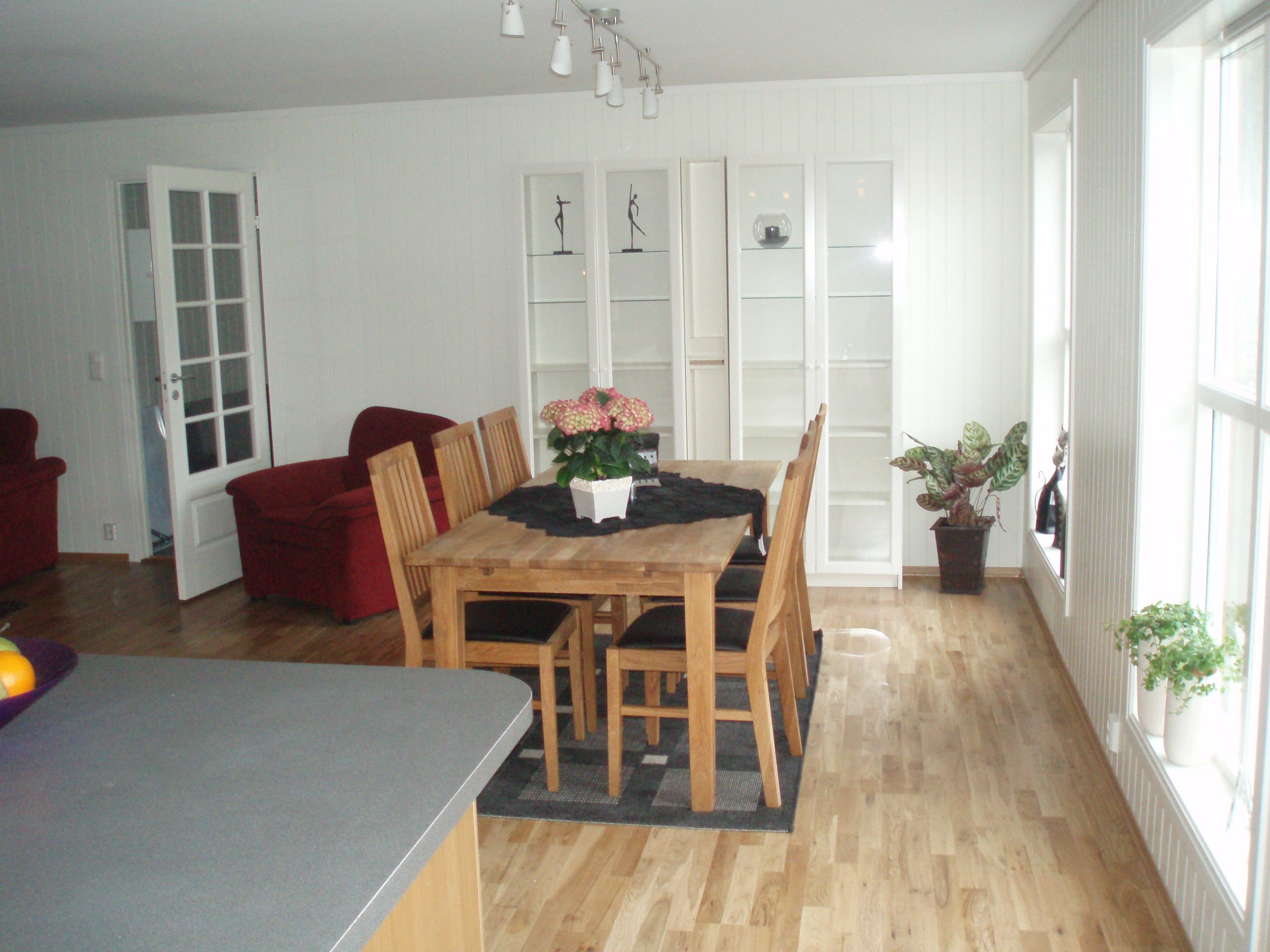 Spisestue. Trebord plass til 6. Røde sofa og sofastol i hjørnet, hvite vitrineskap uten innhold på veggen, og flotte planter på bordet, gulvet, vinduskarmer