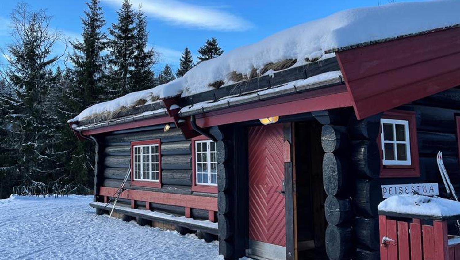Svart og rød hytte i marka. Blå himmel og snø på bakken