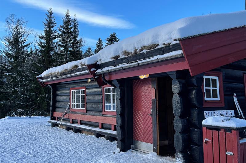 Svart og rød hytte i marka. Blå himmel og snø på bakken