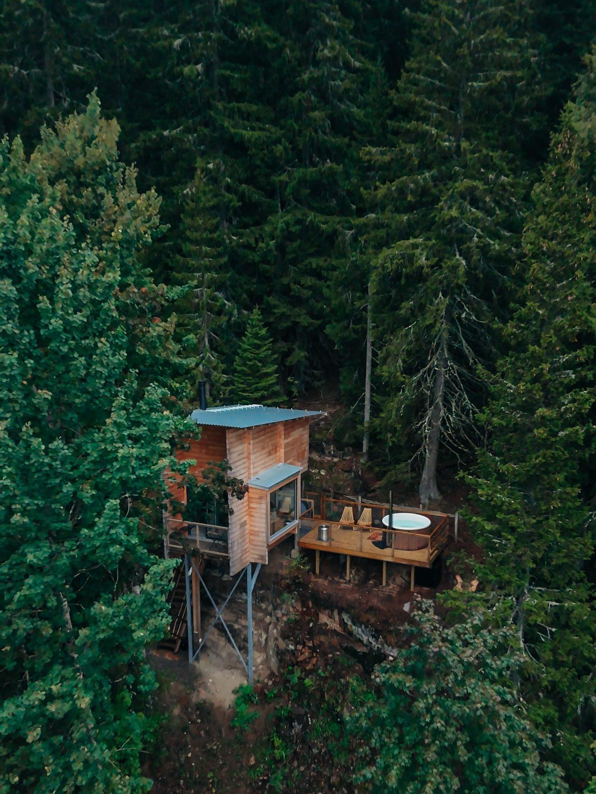 Wooden cabin on stilts surrounded by dense green forest, viewed from above.