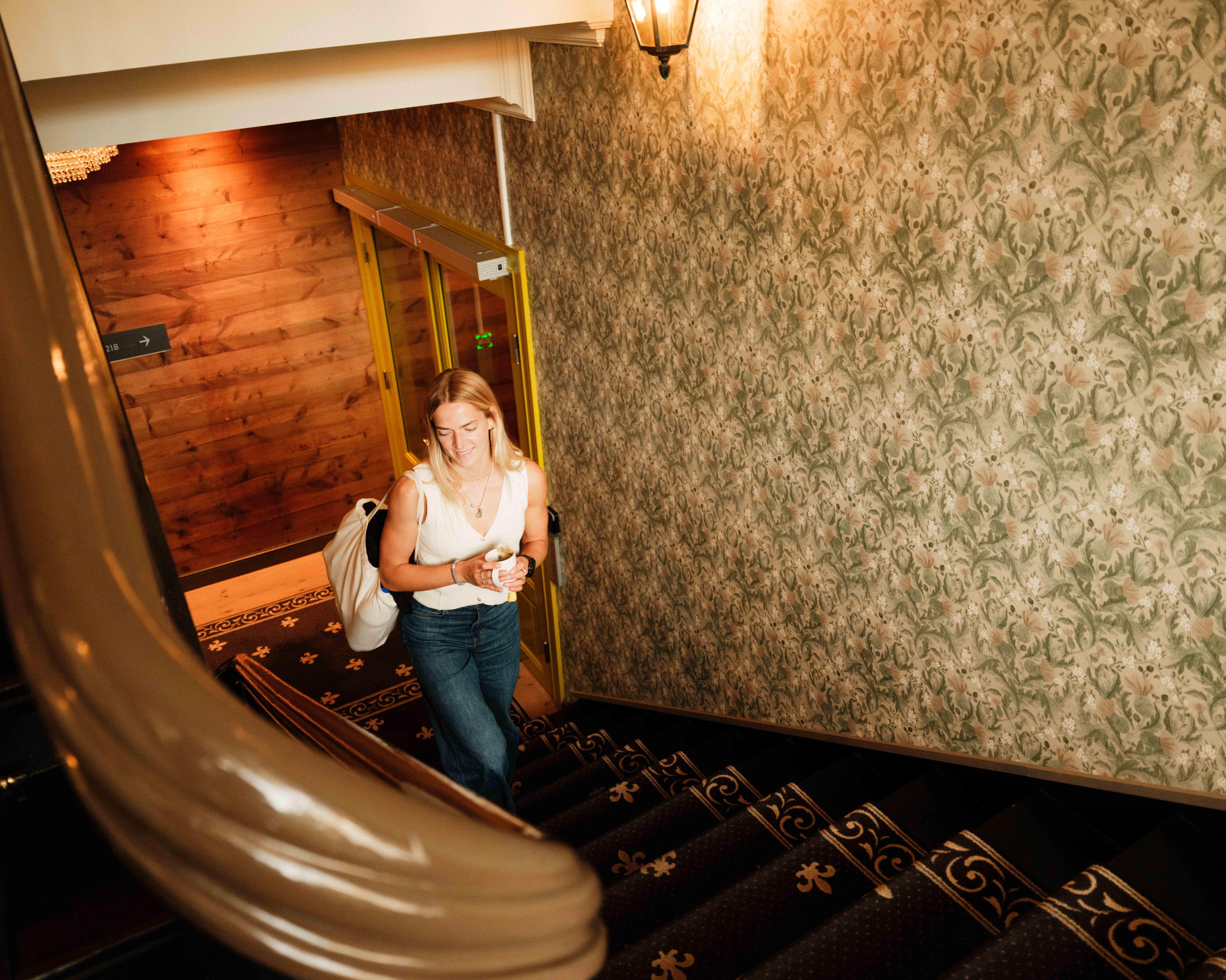 A guest walking up the original staircase at Thon Hotel Sandven – with coffee in hand and bag on her shoulder.