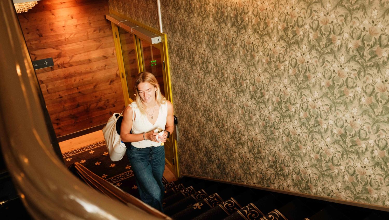 A guest walking up the original staircase at Thon Hotel Sandven – with coffee in hand and bag on her shoulder.