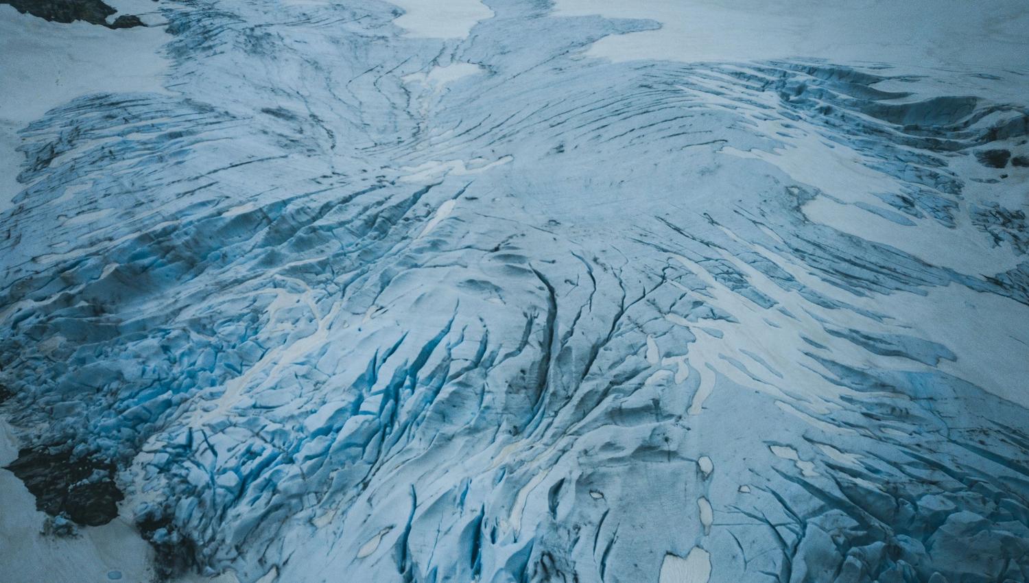 A wide view of a blue-tinged glacier with deep crevasses and snowfields between dark rocky mountains under an overcast sky.