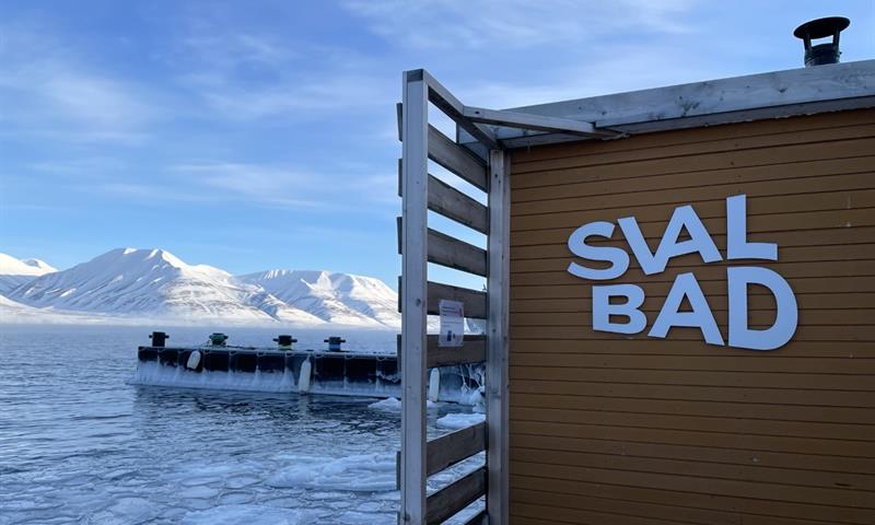 SvalBad's logo on the exterior of the sauna with a fjord and snow-covered mountains in the background