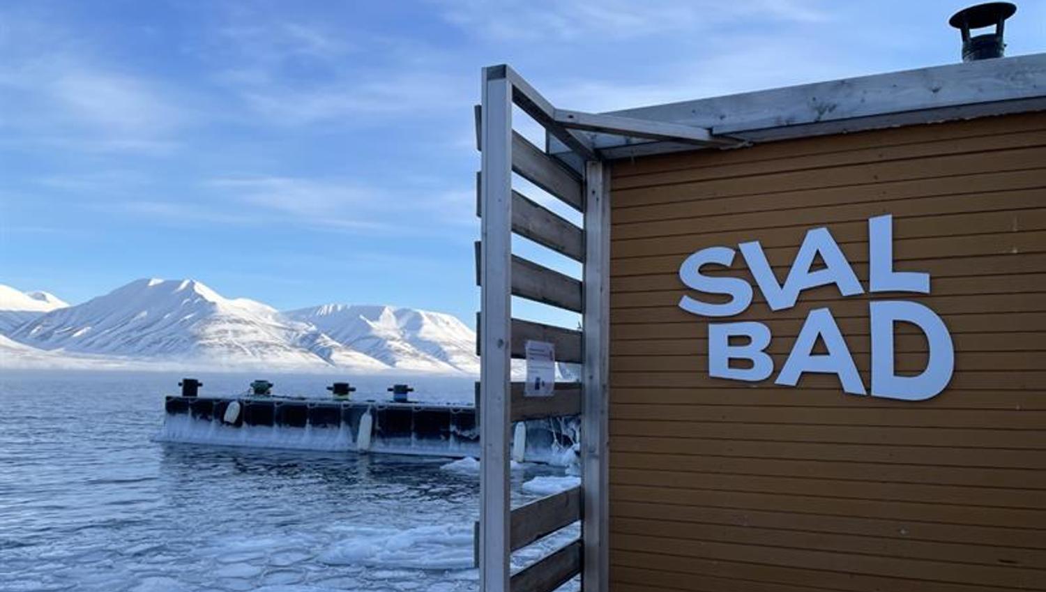 SvalBad's logo on the exterior of the sauna with a fjord and snow-covered mountains in the background