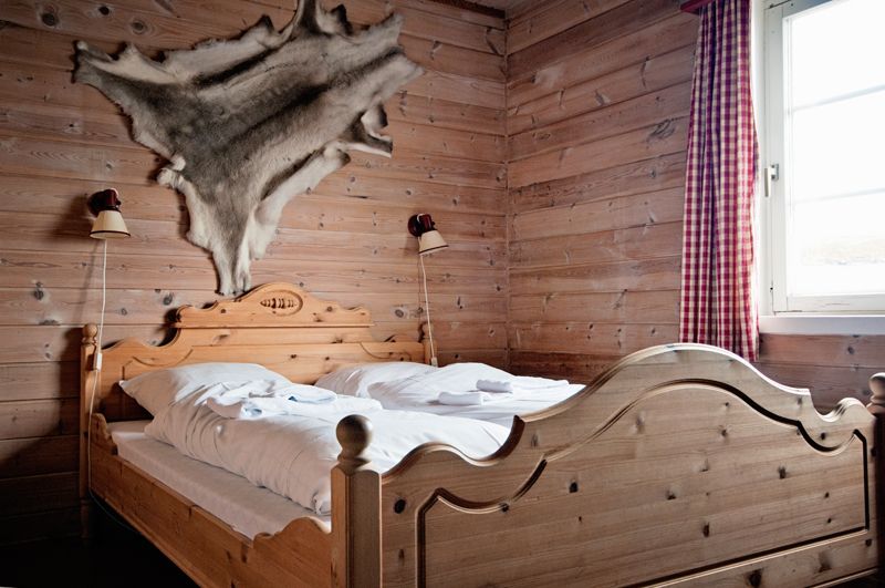 A room with wooden walls, wooden double bed and a reindeer skin hanging on the wall above the bed.