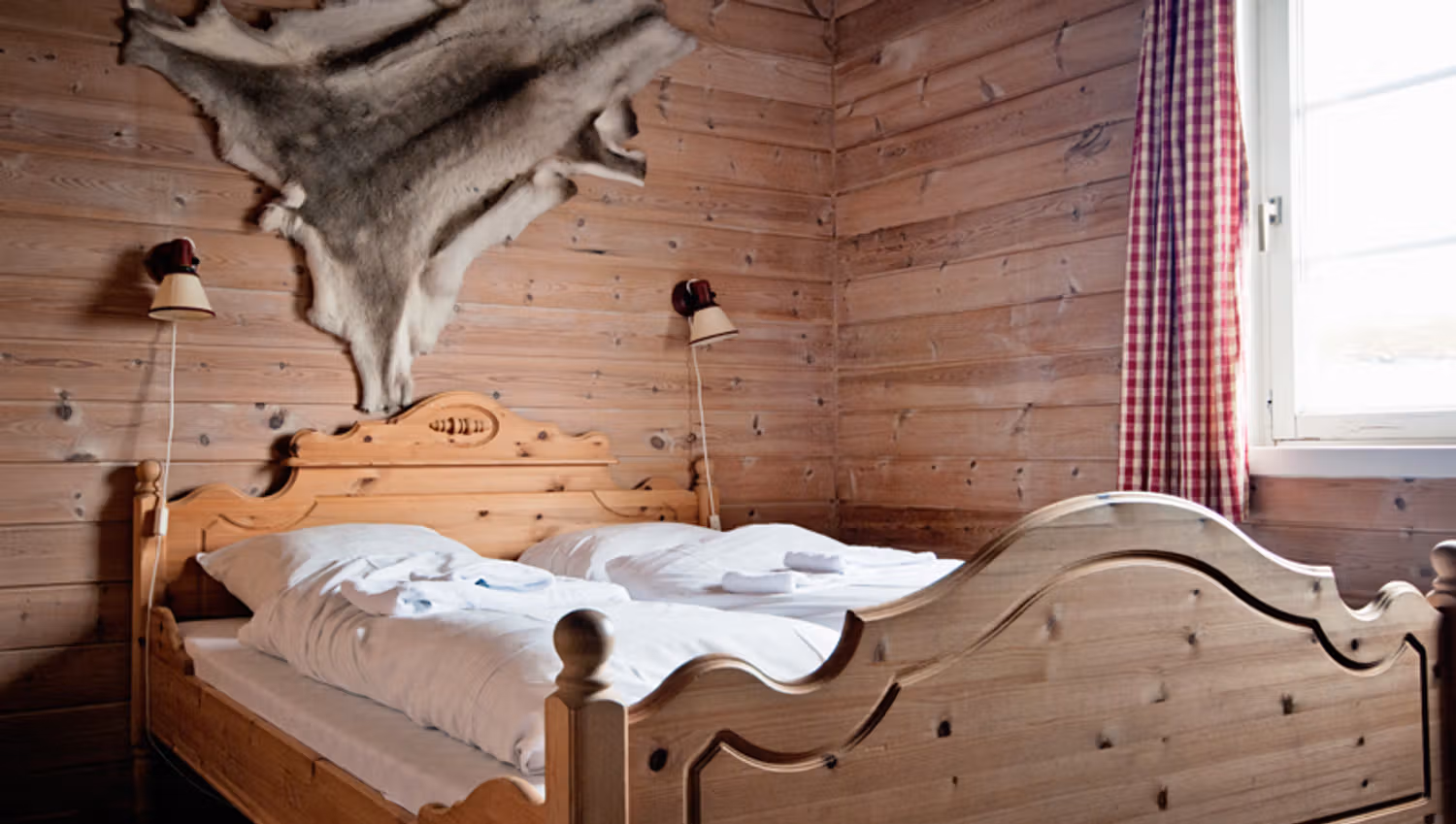 A room with wooden walls, wooden double bed and a reindeer skin hanging on the wall above the bed.