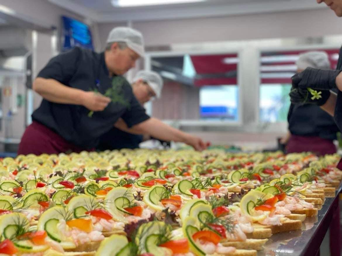 A kitchen staff member prepares a long table of open sandwiches decorated with vegetables and shrimp.