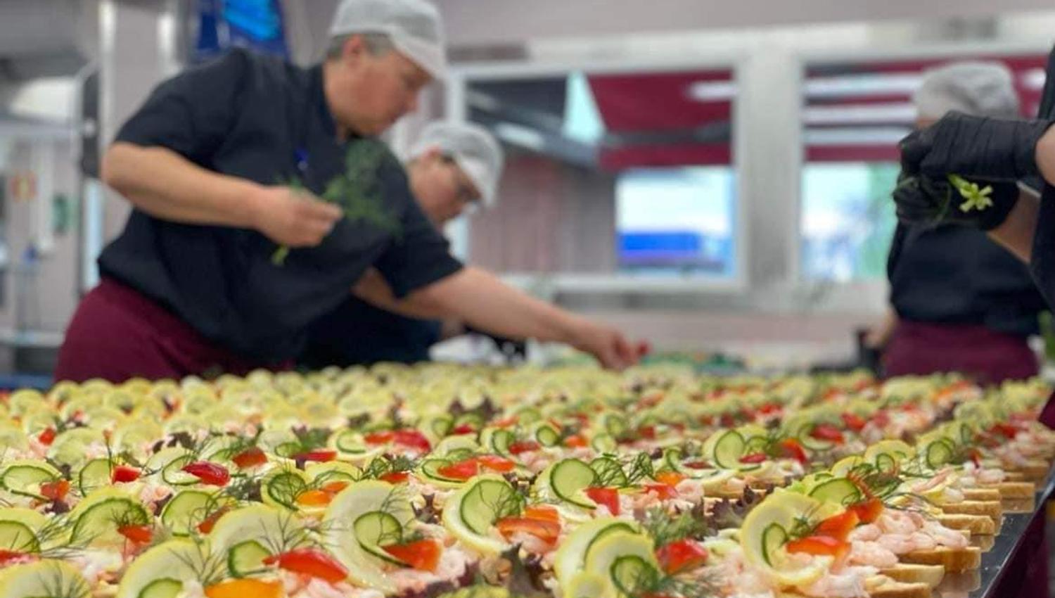 A kitchen staff member prepares a long table of open sandwiches decorated with vegetables and shrimp.