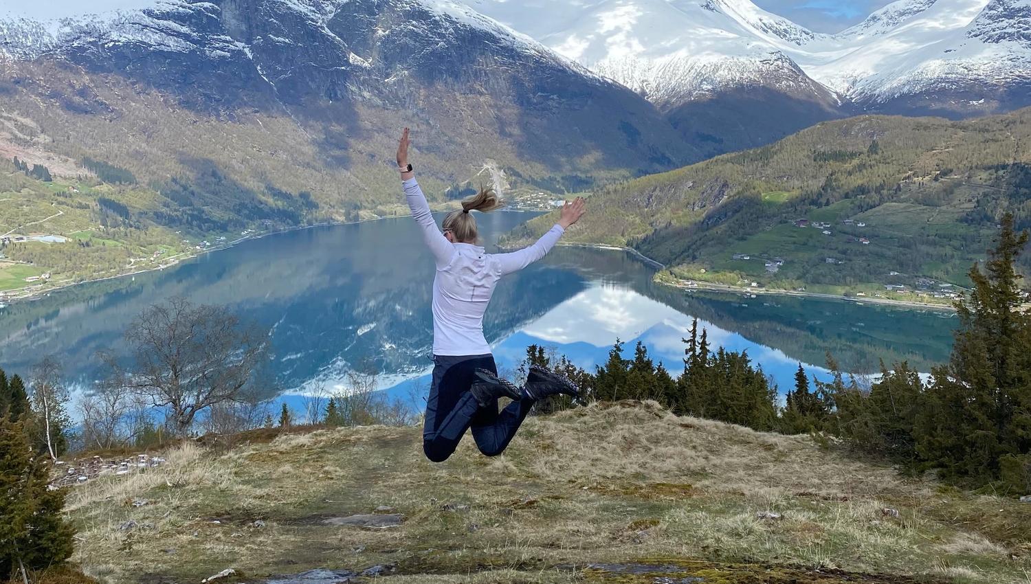 Person mid-jump on a grassy hill overlooking a blue fjord, forested slopes, and snow-capped mountains under clouds at Skarsteinssætra in Olden.