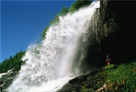 Powerful waterfall pours over a rocky cliff into mist, with a small hiker standing on green vegetation below.