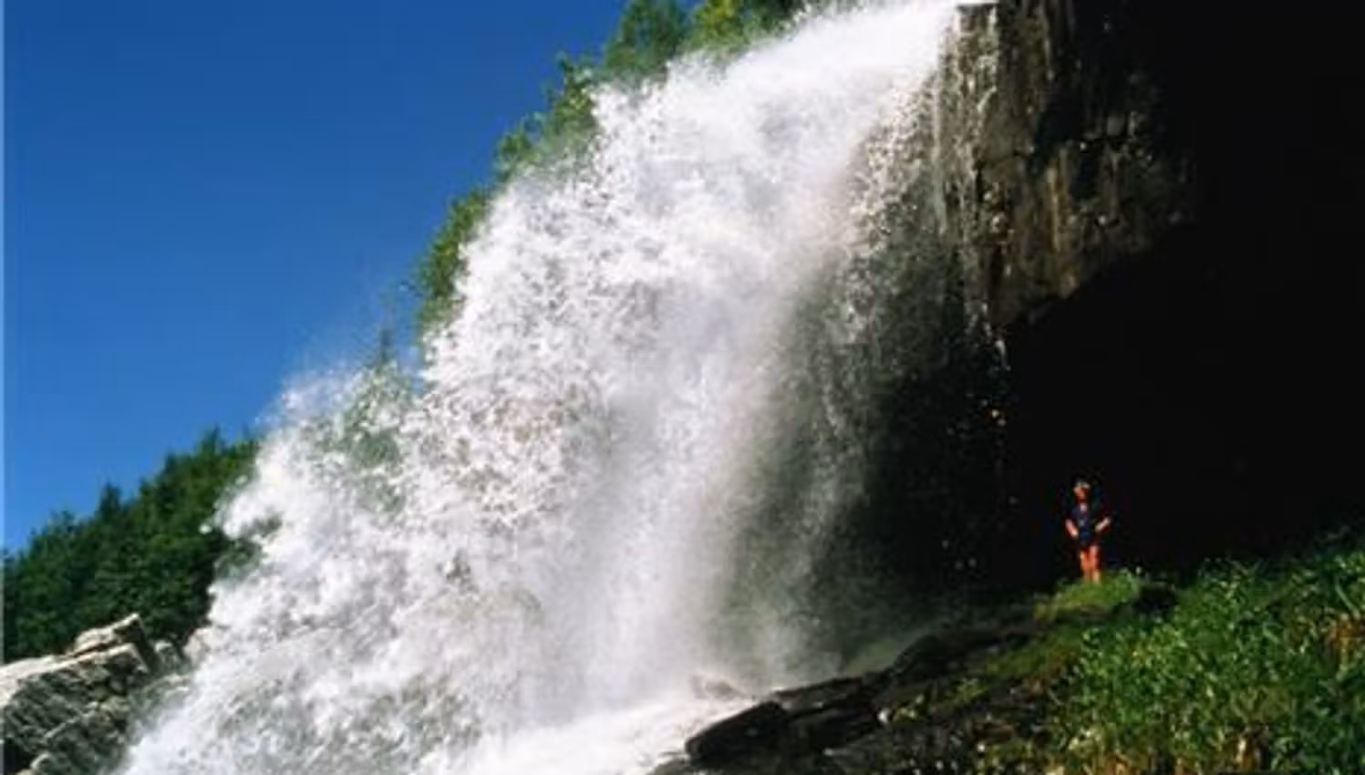 Powerful waterfall pours over a rocky cliff into mist, with a small hiker standing on green vegetation below.