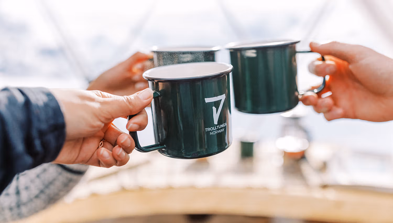 Two individuals clinking green enamel mugs labeled "Trolltunga Norway" in a cozy outdoor setting.