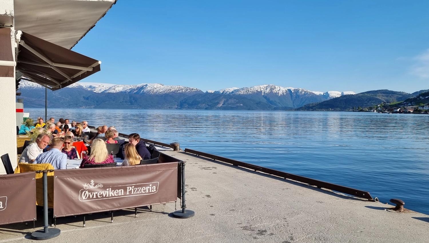 Guests enjoying food in the sun at Øvreviken Pizzeria’s outdoor seating by the Hardangerfjord, overlooking snowy mountains.