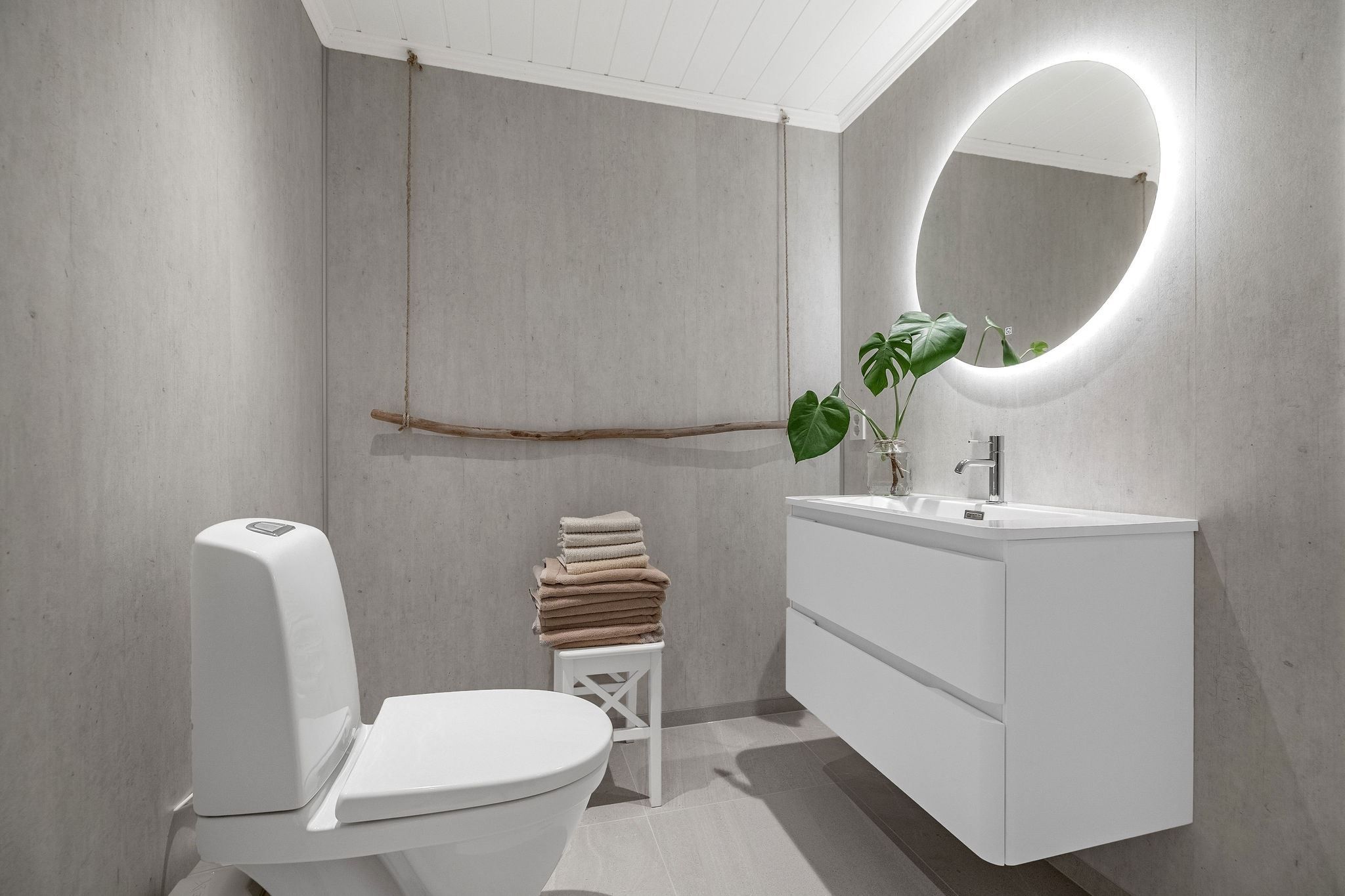 Minimalist bathroom with a white toilet, floating vanity and sink, round lit mirror, towels on stool, and plant at the Seahouse at Hornelen Panorama in Bremanger..