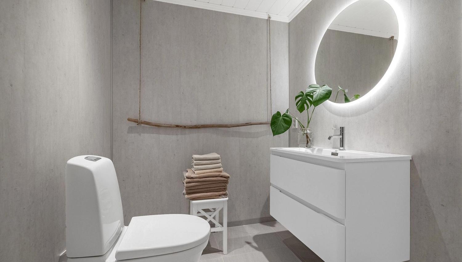 Minimalist bathroom with a white toilet, floating vanity and sink, round lit mirror, towels on stool, and plant at the Seahouse at Hornelen Panorama in Bremanger..