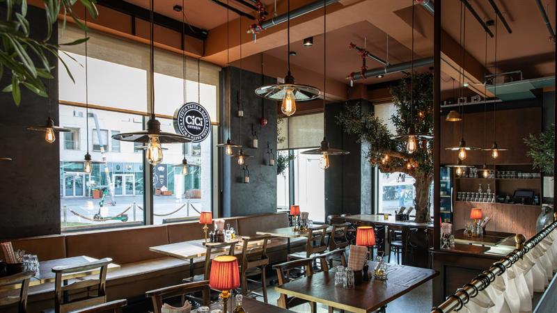 Restaurantinteriør med store vinduer, brun skinnsofa langs veggen, trestoler, bordlamper med røde skjermer og hengende industrielle lamper