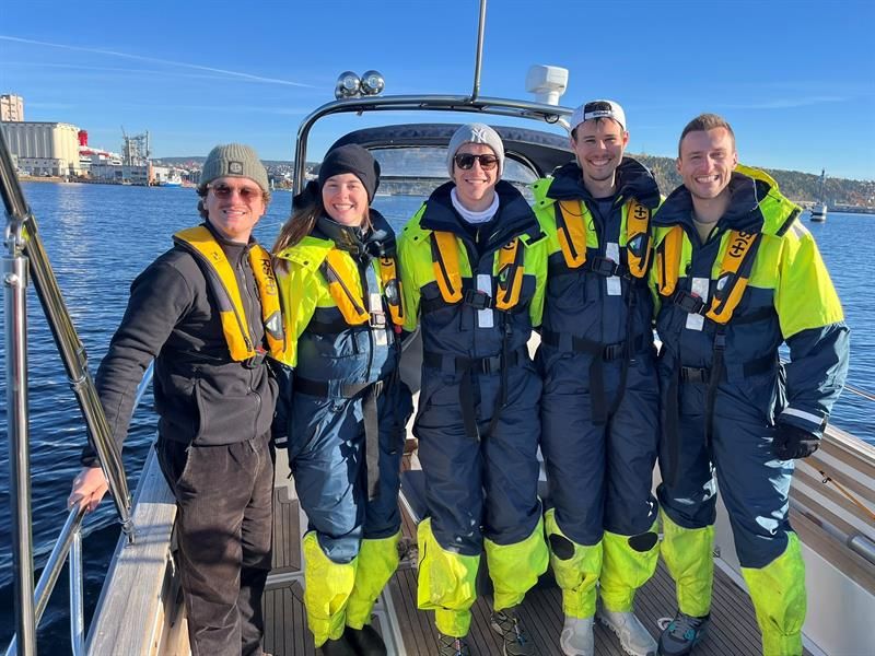 A group of smiling people in survival suits stand together on a boat in the sunshine.