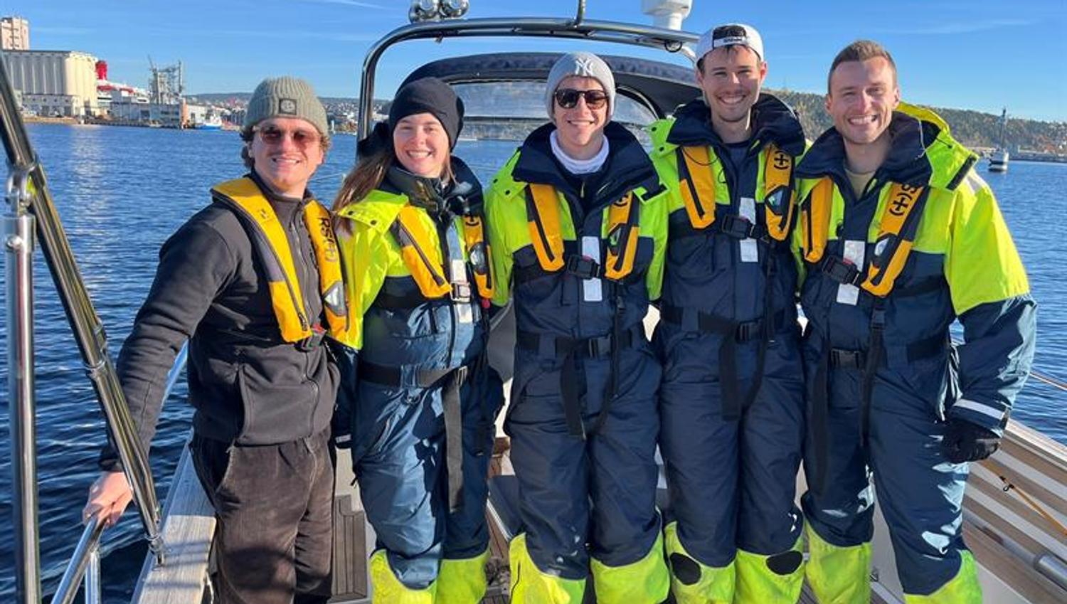 A group of smiling people in survival suits stand together on a boat in the sunshine.
