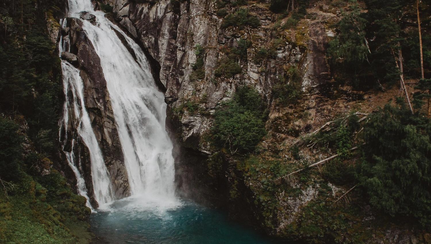 A tall waterfall cascades down a rocky cliff into a turquoise pool, surrounded by forested slopes and mossy grass.
