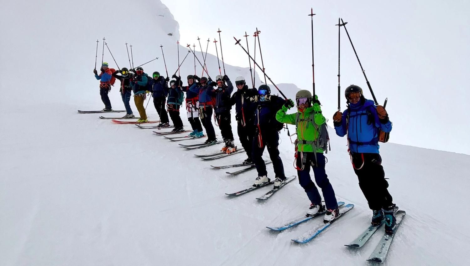 A line of skiers on a snowy mountain ridge raise ski poles, wearing helmets, goggles, and backpacks in foggy light..