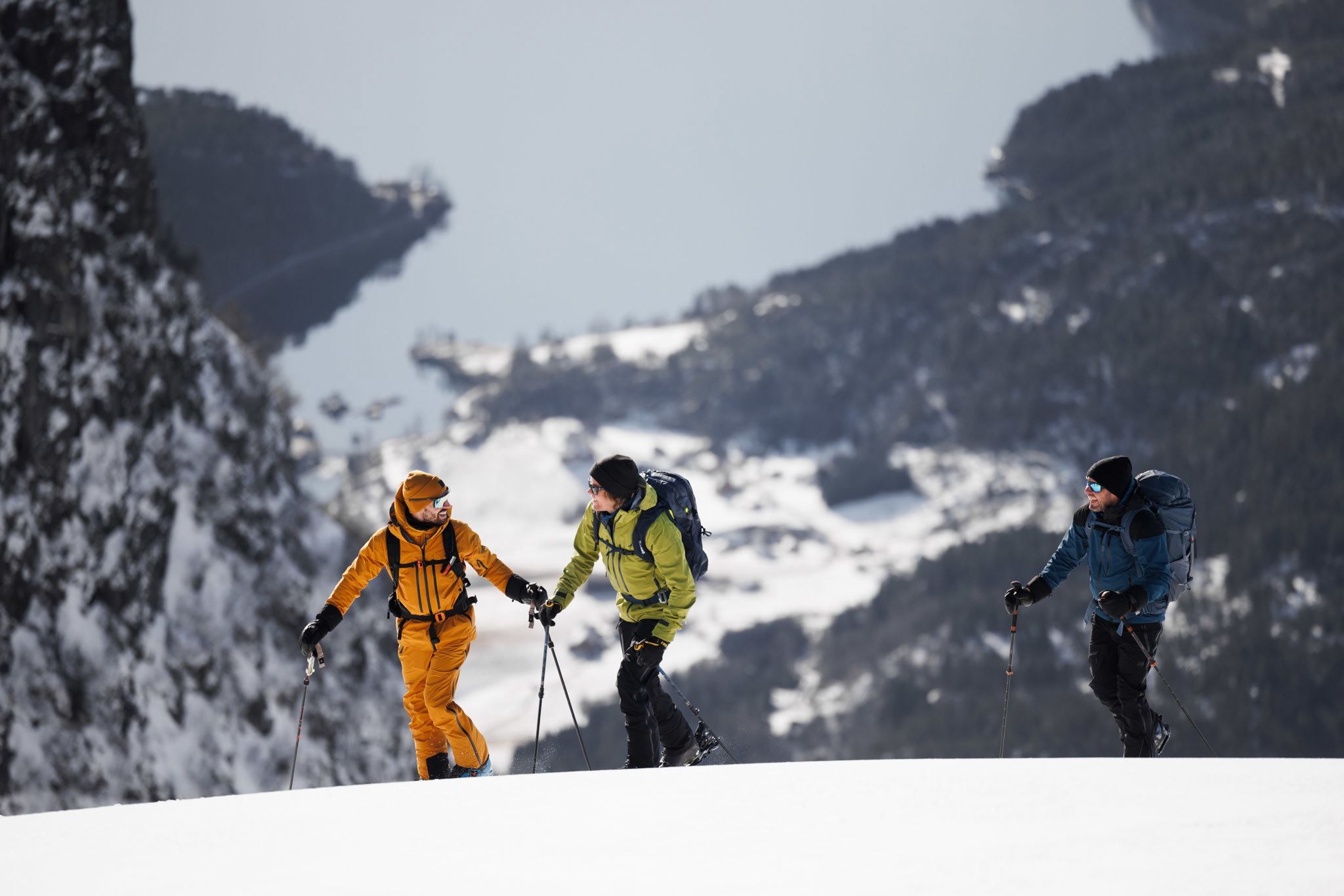 Three hikers with backpacks and trekking poles walk along a snow-covered ridge with distant mountains behind.