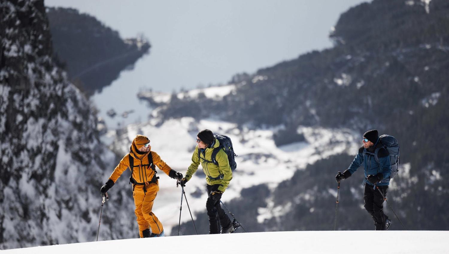 Three hikers with backpacks and trekking poles walk along a snow-covered ridge with distant mountains behind.