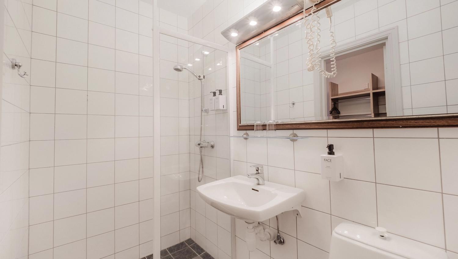 White tiled bathroom with a glass shower stall, wall-mounted sink, large mirror, and toilet in a bright interior.