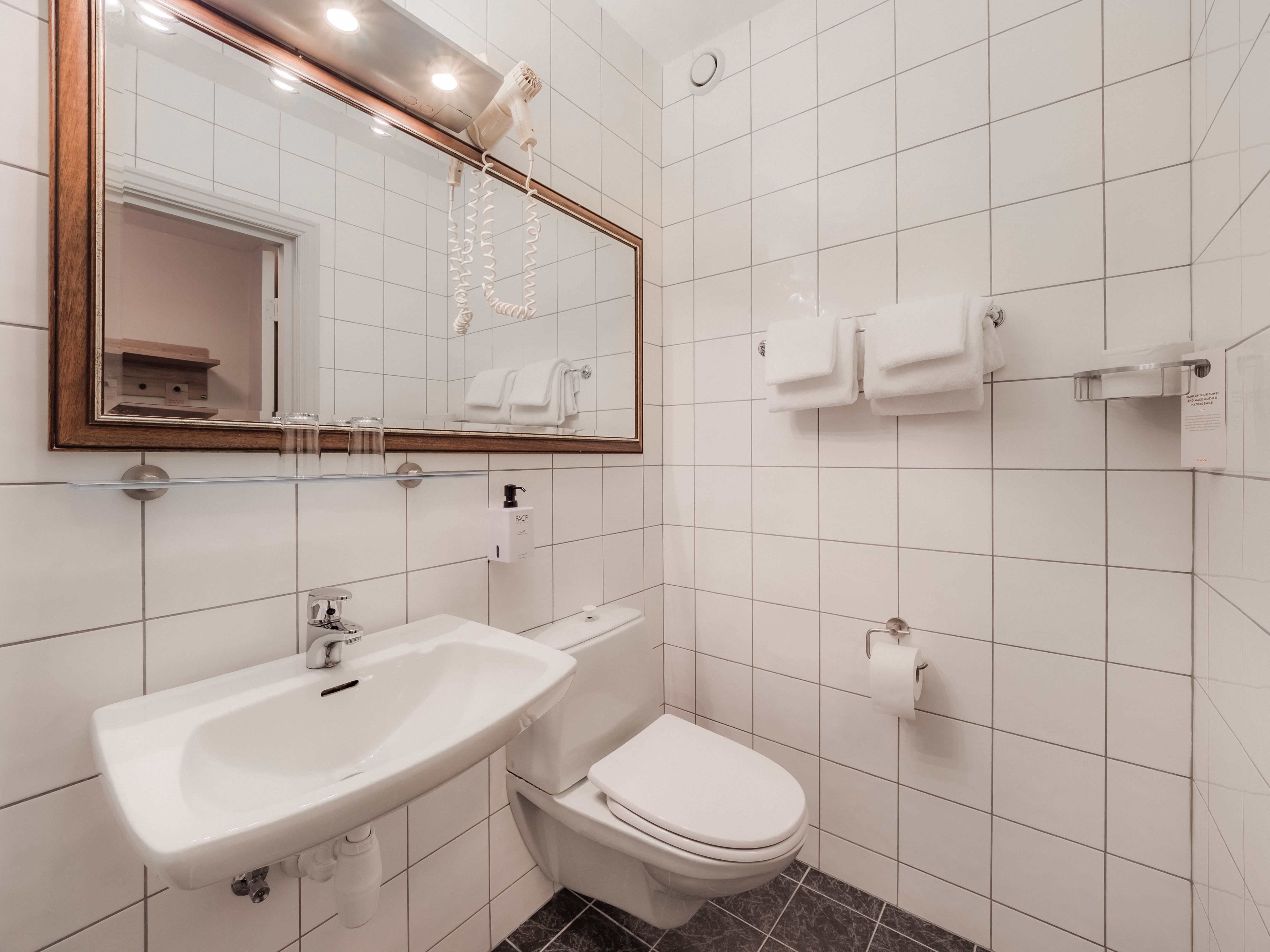 Clean bathroom with white tiled walls, wall-mounted sink, toilet, large mirror, towels, and a hair dryer.