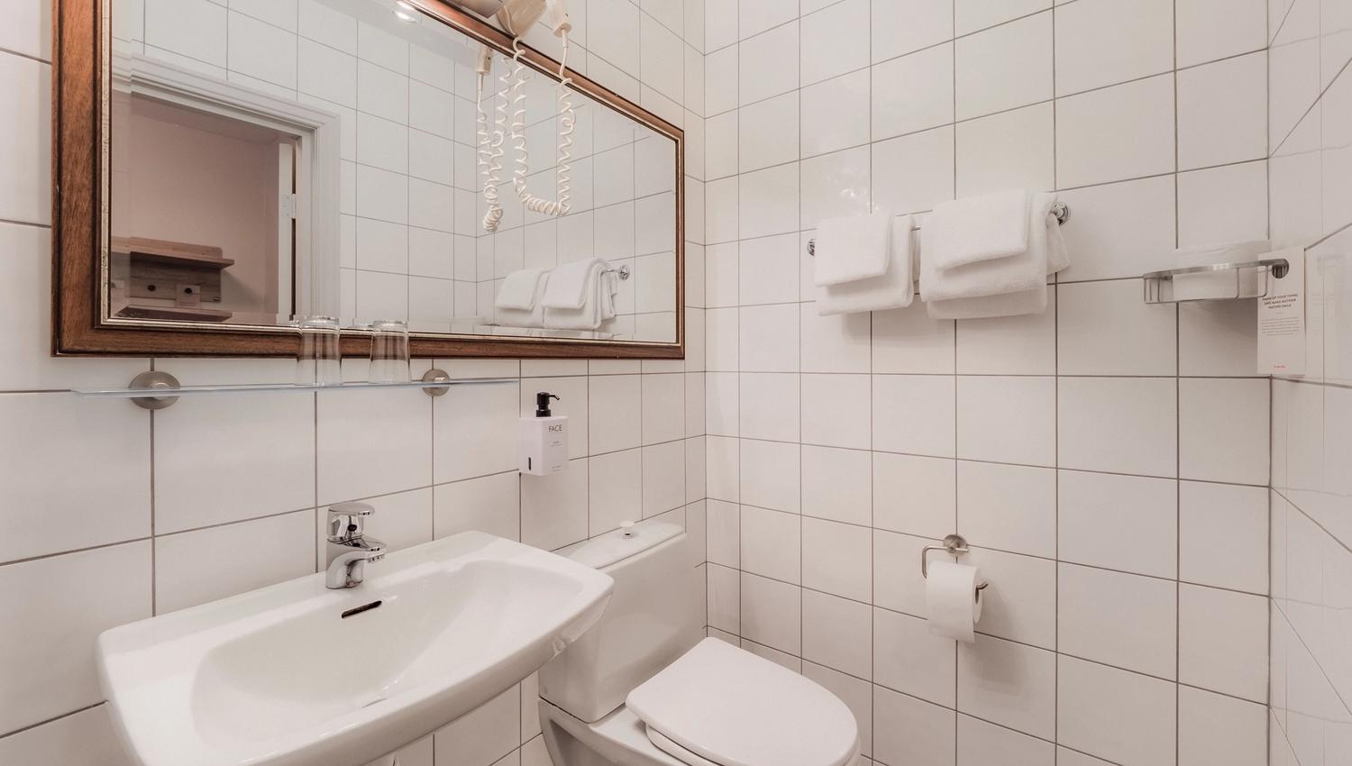 Clean bathroom with white tiled walls, wall-mounted sink, toilet, large mirror, towels, and a hair dryer.