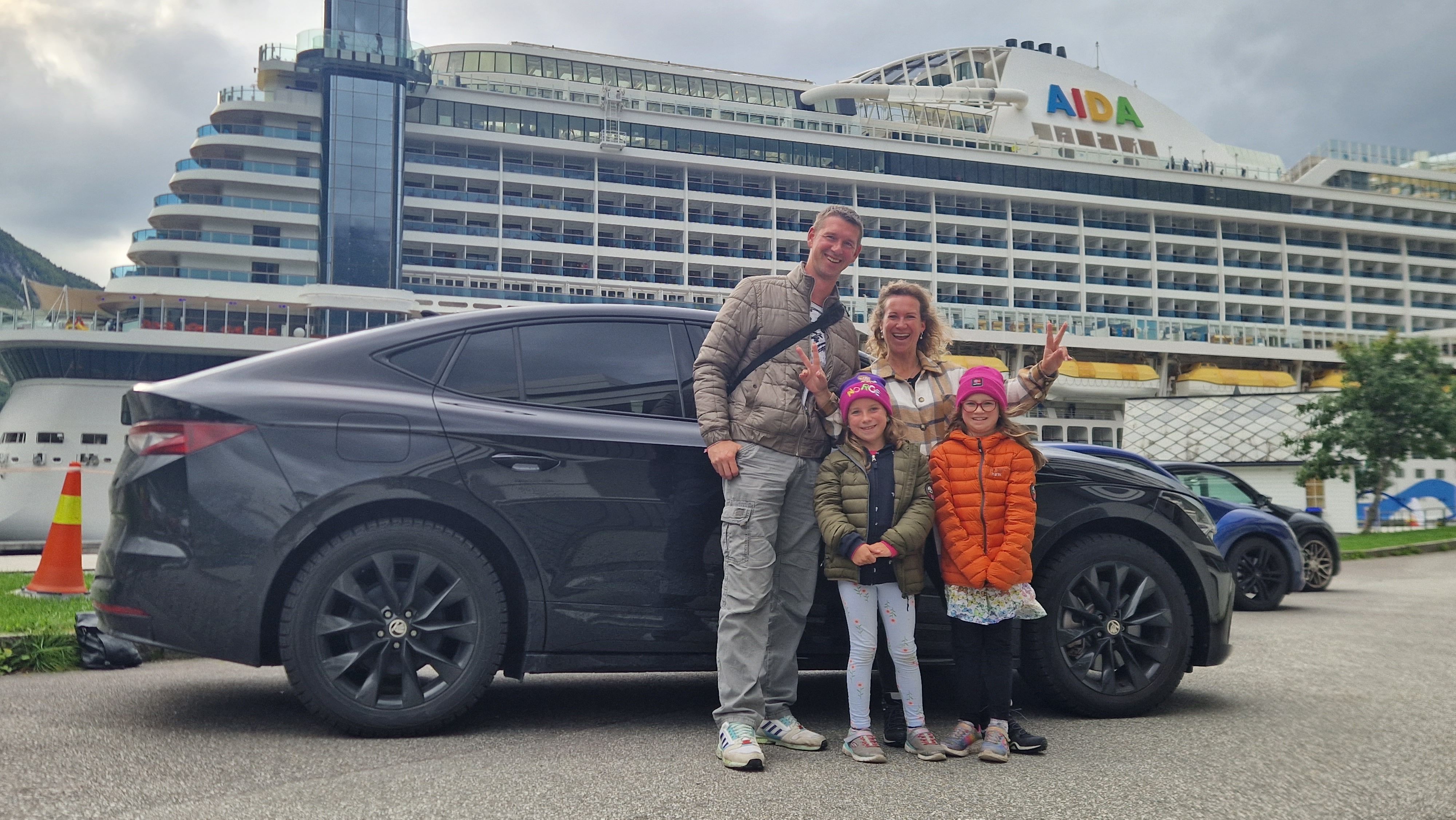 A family of four standing in front of a car. There is a cruise ship in the background. 