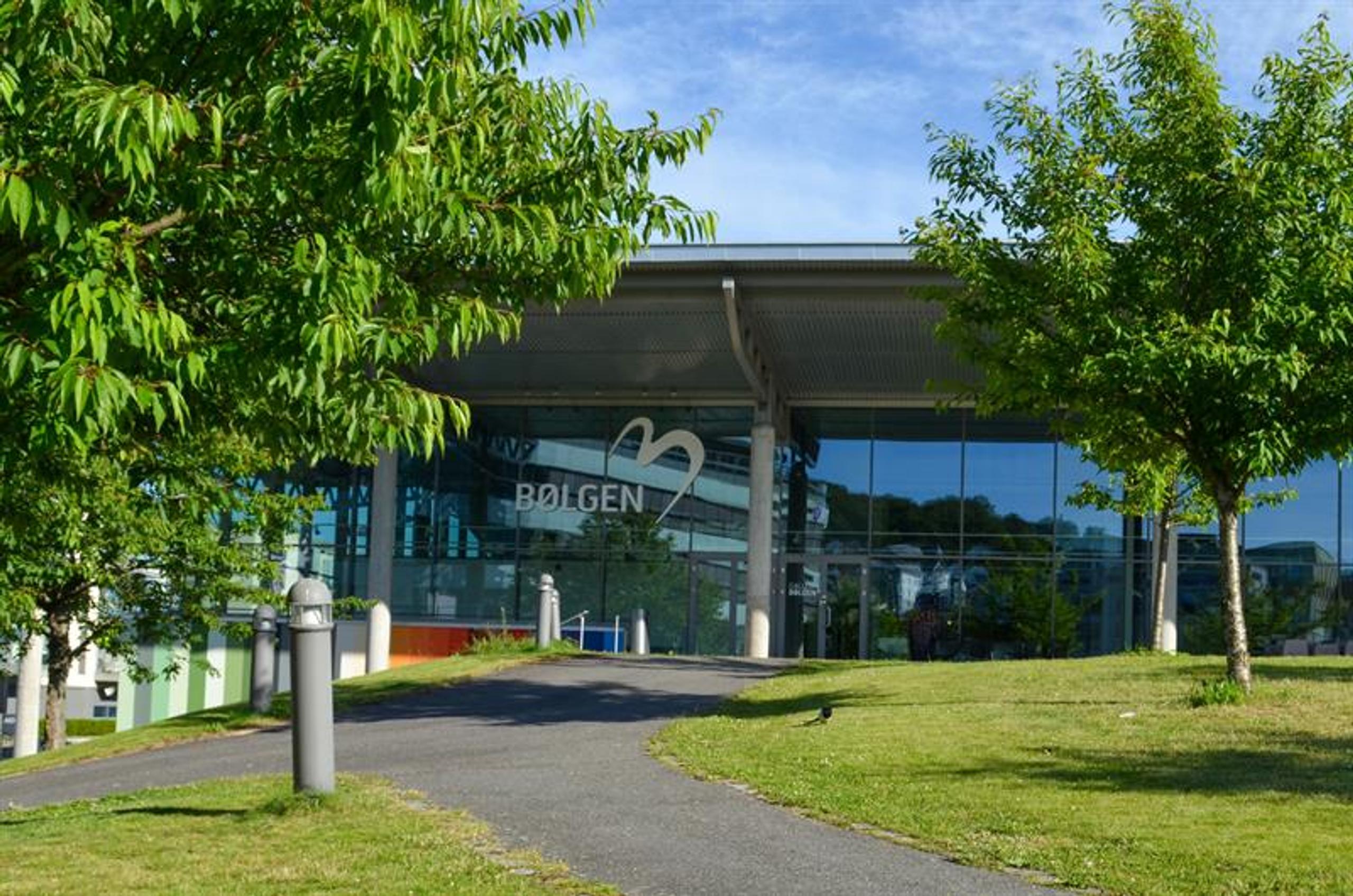 Bølgen Cultural Centre in Larvik, featuring a modern glass façade, a walkway and green trees in front of the building on a sunny day.