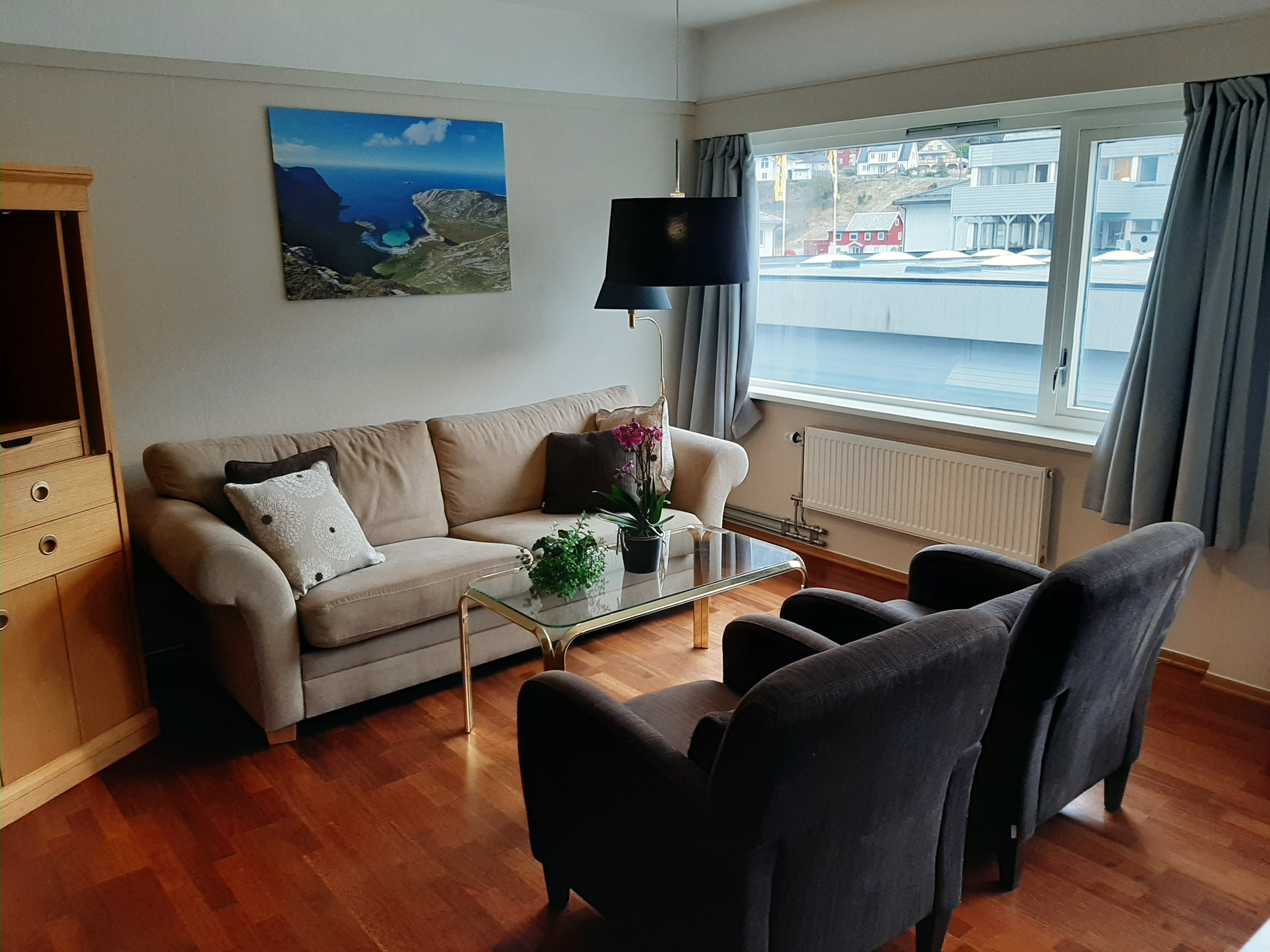 Ei lys stove med beige sofa, to mørke lenestolar, glasbord med plantar og stort vindauge med gardiner på Bremanger Fjordhotell..