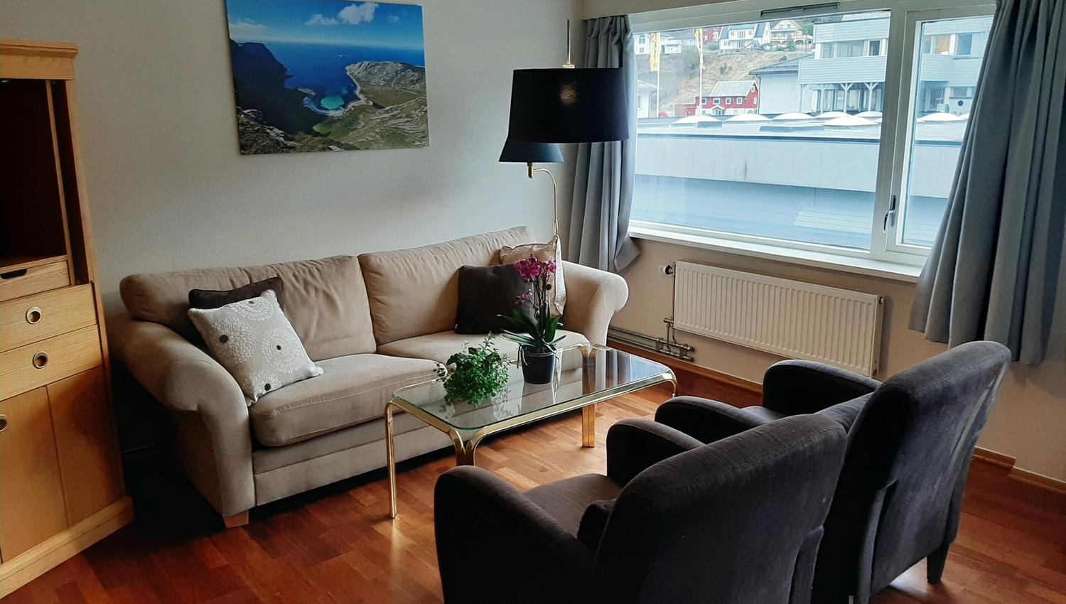 Ei lys stove med beige sofa, to mørke lenestolar, glasbord med plantar og stort vindauge med gardiner på Bremanger Fjordhotell..