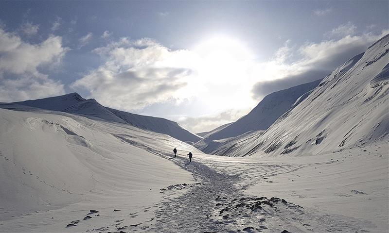 To personer som går opp et fjell langs en snødekt sti i et snøfylt fjellandskap med solskinn og en lett skyet himmel i bakgrunnen