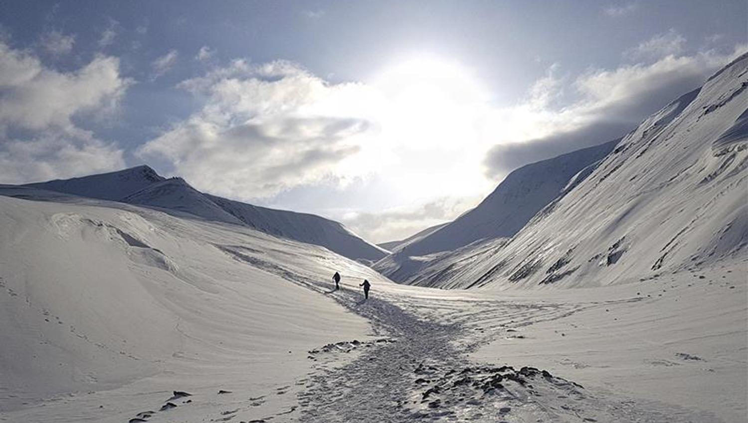 To personer som går opp et fjell langs en snødekt sti i et snøfylt fjellandskap med solskinn og en lett skyet himmel i bakgrunnen