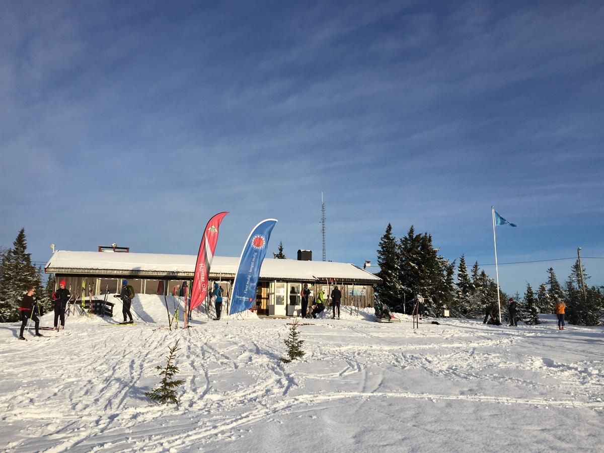 A winter cabin or mountain lodge surrounded by snow-covered terrain, with flags and banner flags standing in front of the building under a clear sky.