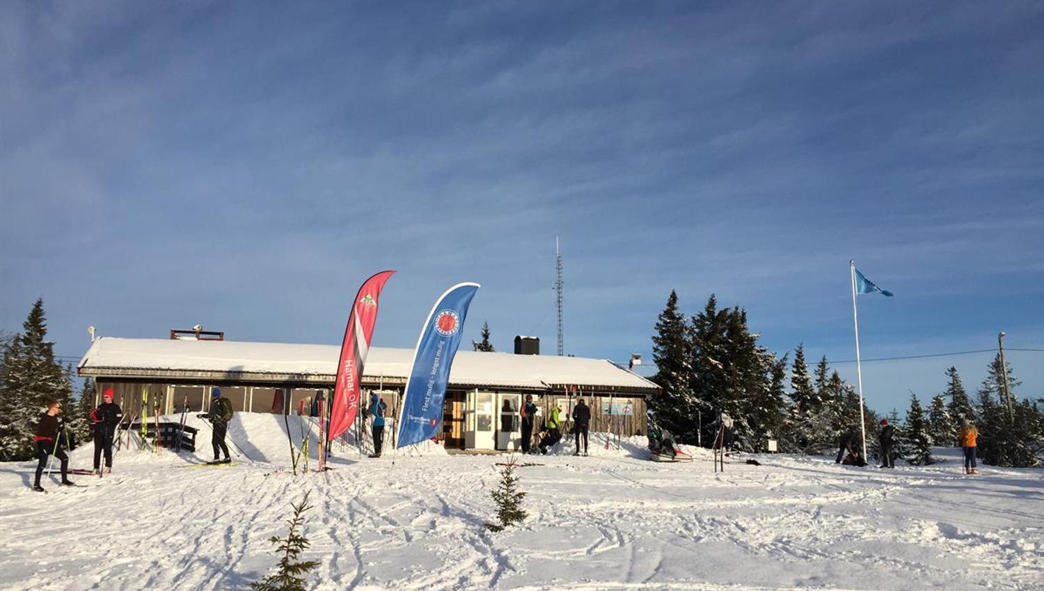 A winter cabin or mountain lodge surrounded by snow-covered terrain, with flags and banner flags standing in front of the building under a clear sky.