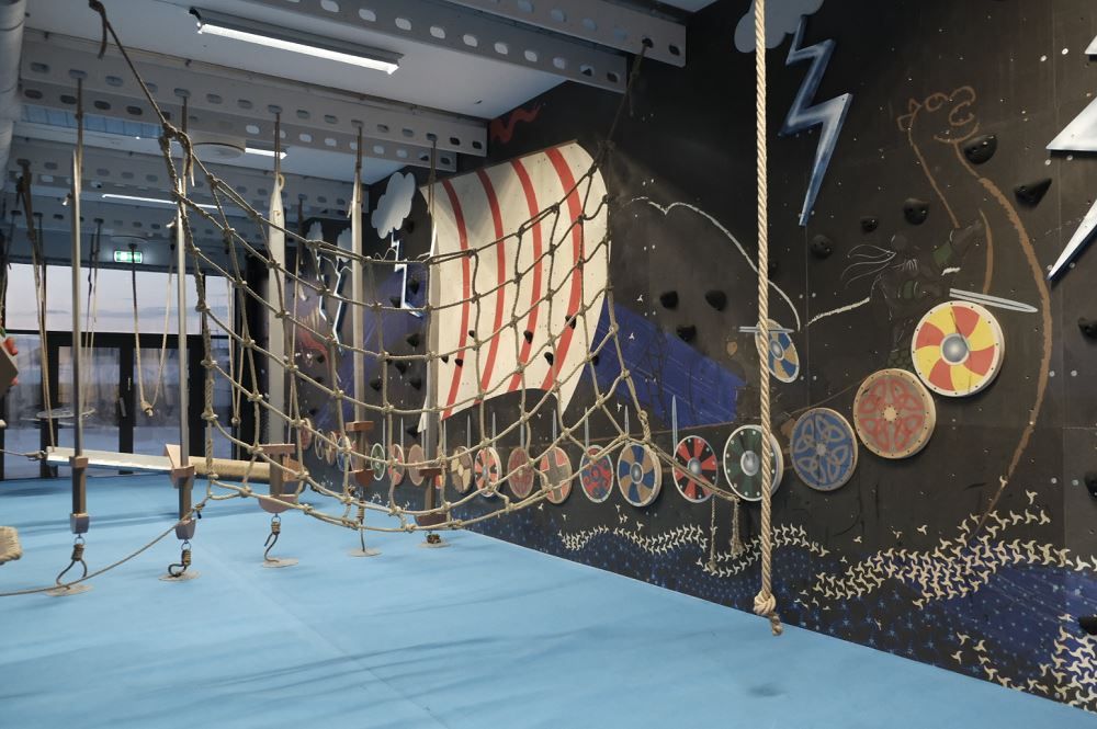 Indoor adventure course with rope net bridge, hanging steps, blue padded floor, and Viking-themed mural wall..