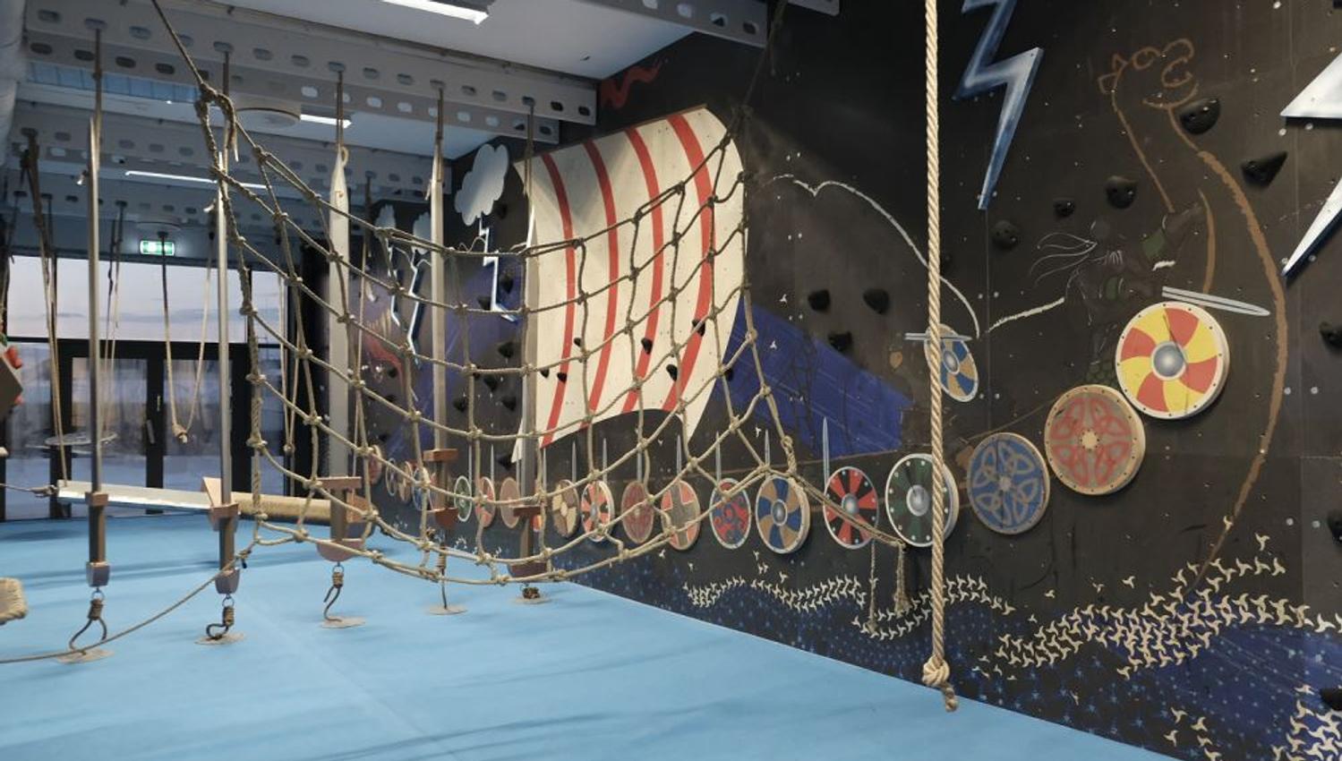 Indoor adventure course with rope net bridge, hanging steps, blue padded floor, and Viking-themed mural wall..