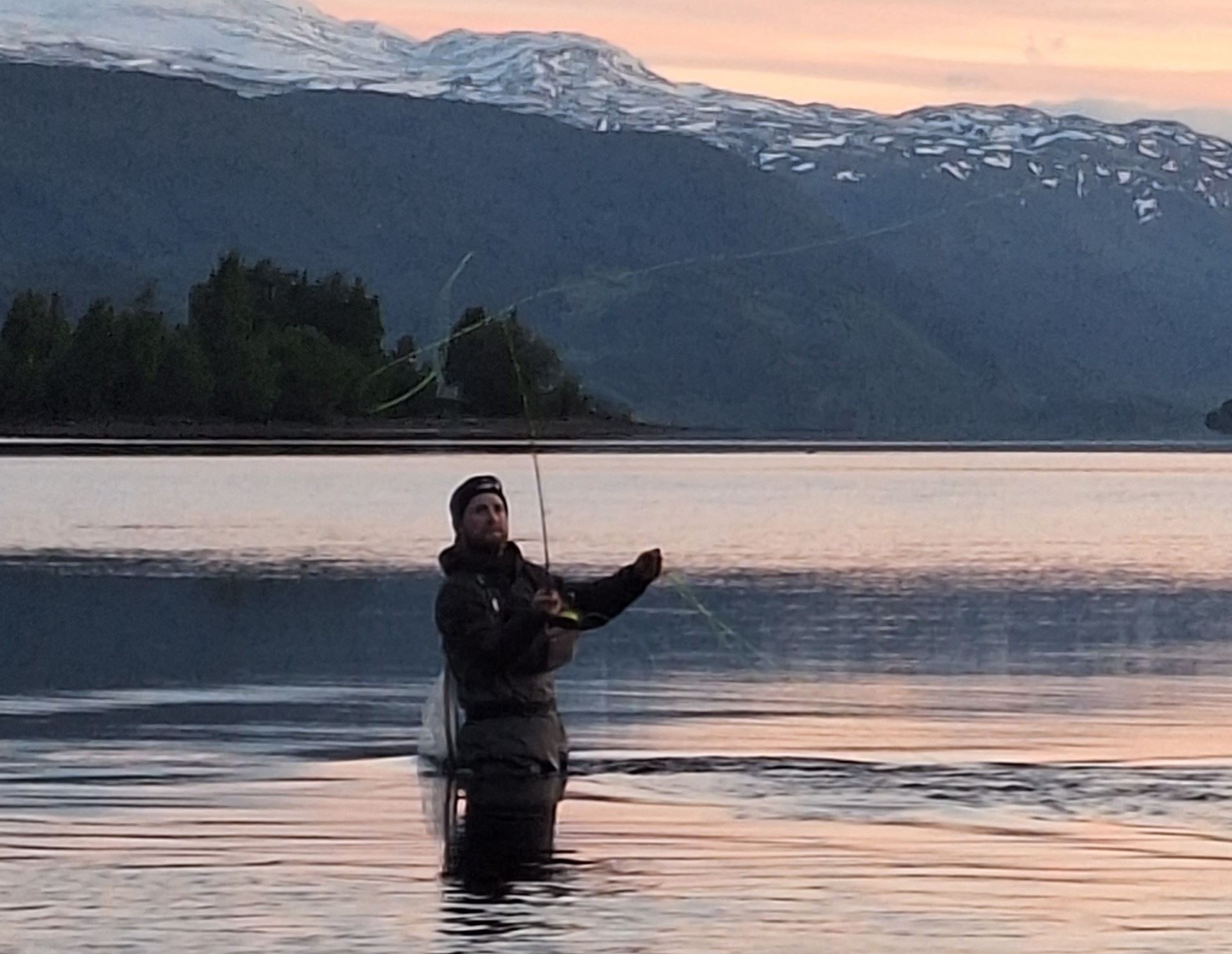 Person står i vann og fisker i innsjø i skumring, med fjell i bakgrunnen.