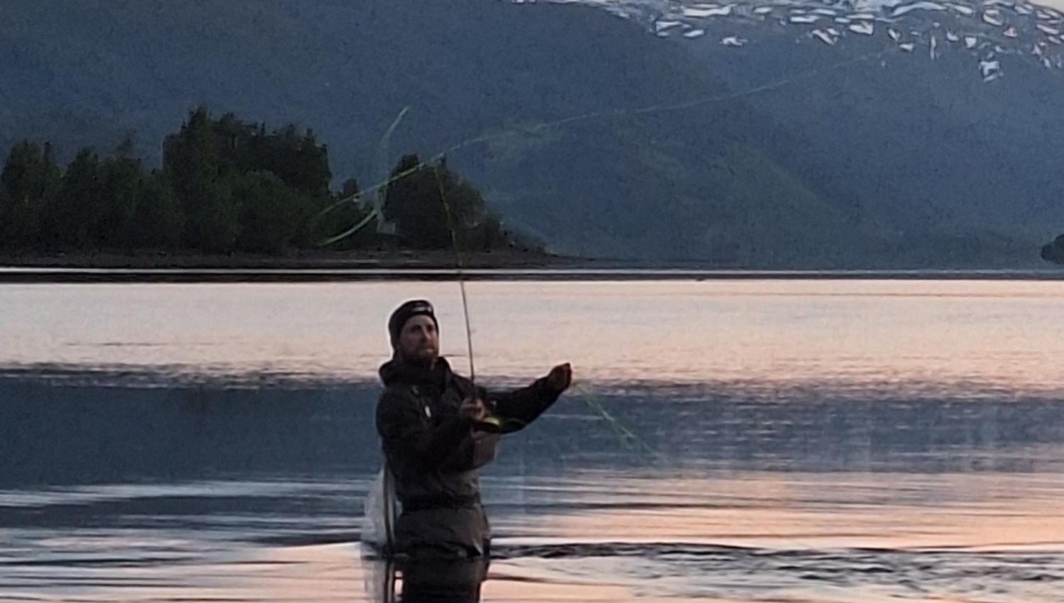 Person står i vann og fisker i innsjø i skumring, med fjell i bakgrunnen.
