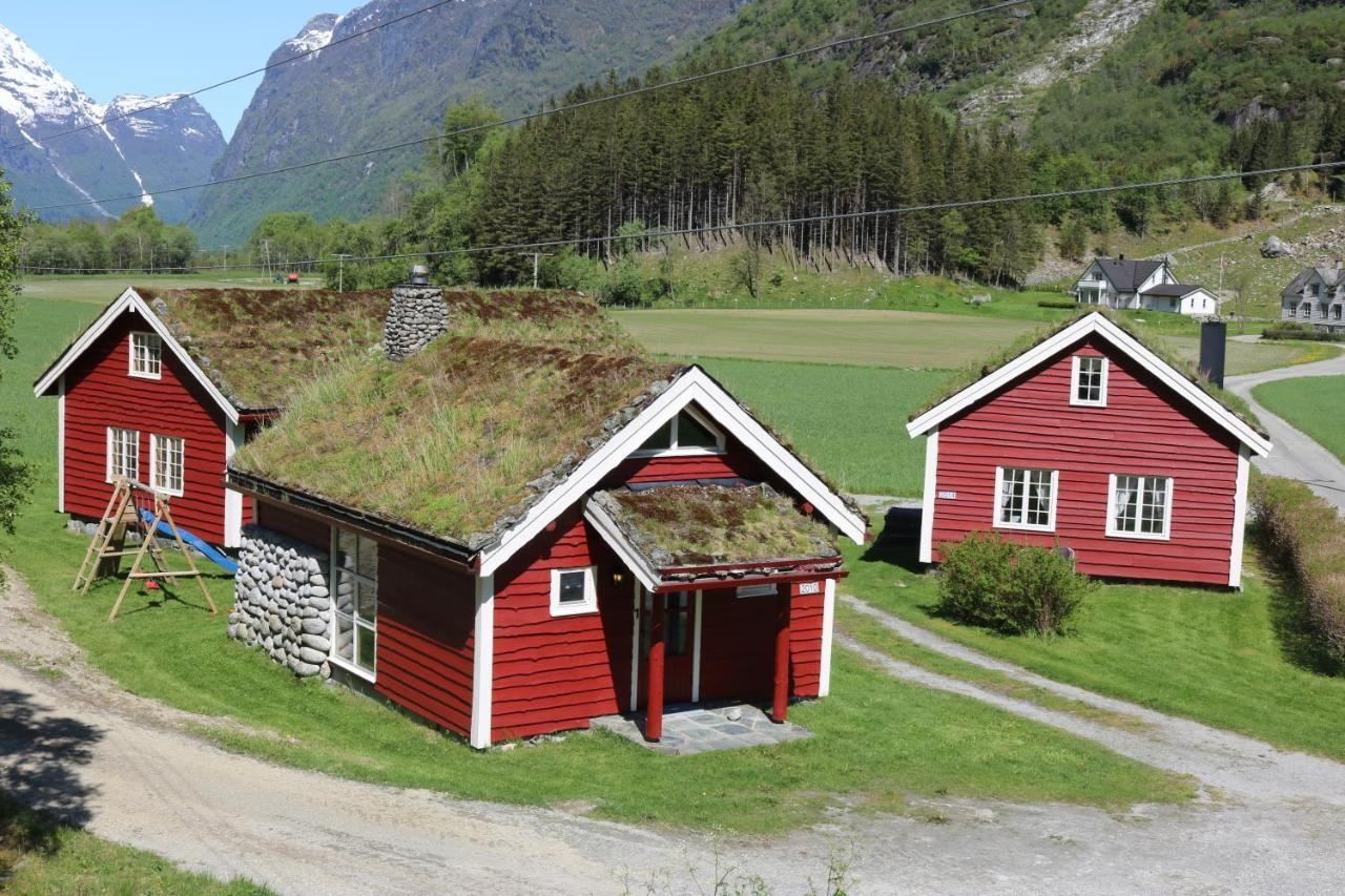 Three red wooden houses with grass roofs sit on green fields, with forested hills and snowy mountains behind, at Aabrekk Gard Trollbu in Oldedalen valley.