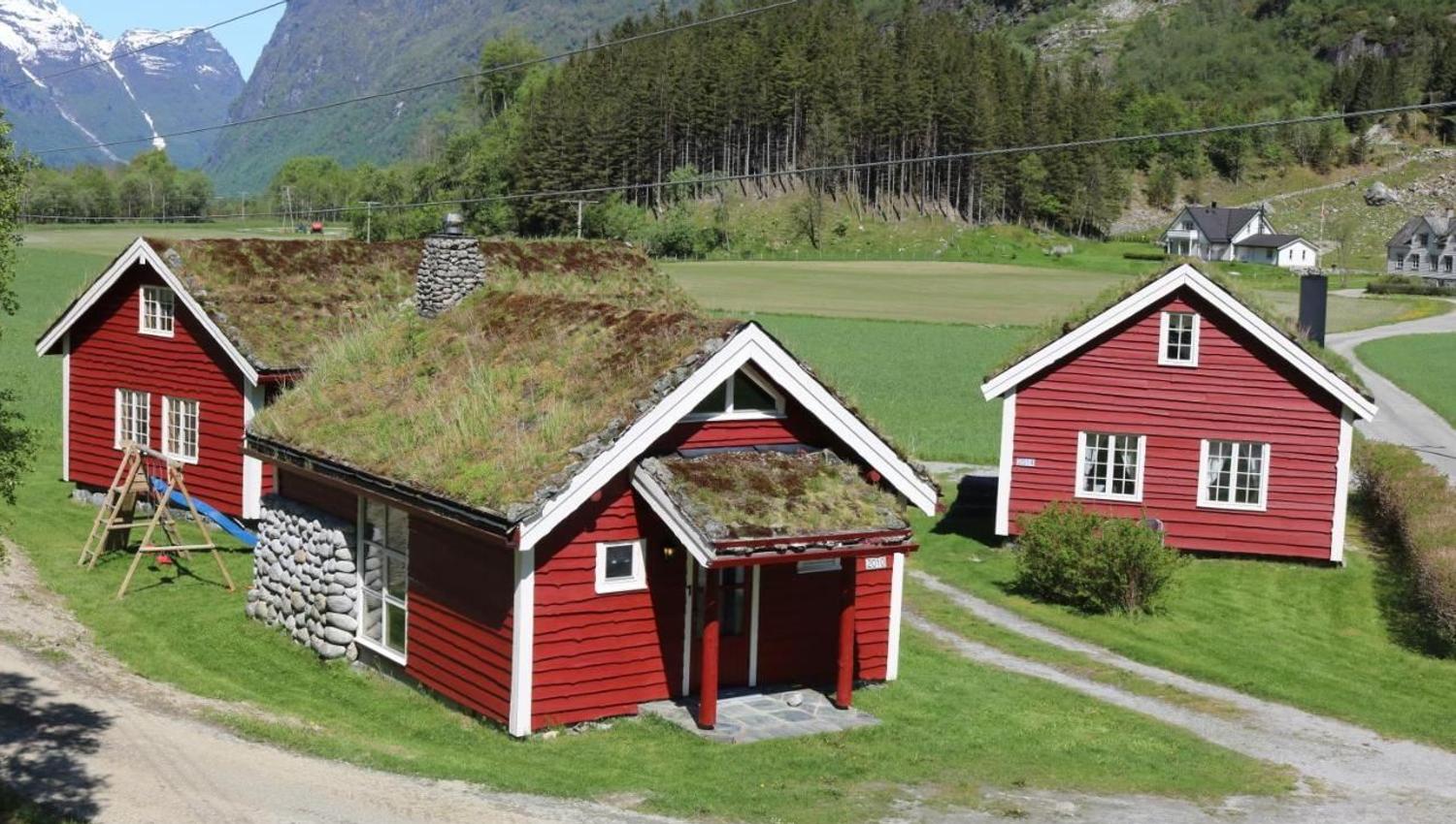 Three red wooden houses with grass roofs sit on green fields, with forested hills and snowy mountains behind, at Aabrekk Gard Trollbu in Oldedalen valley.