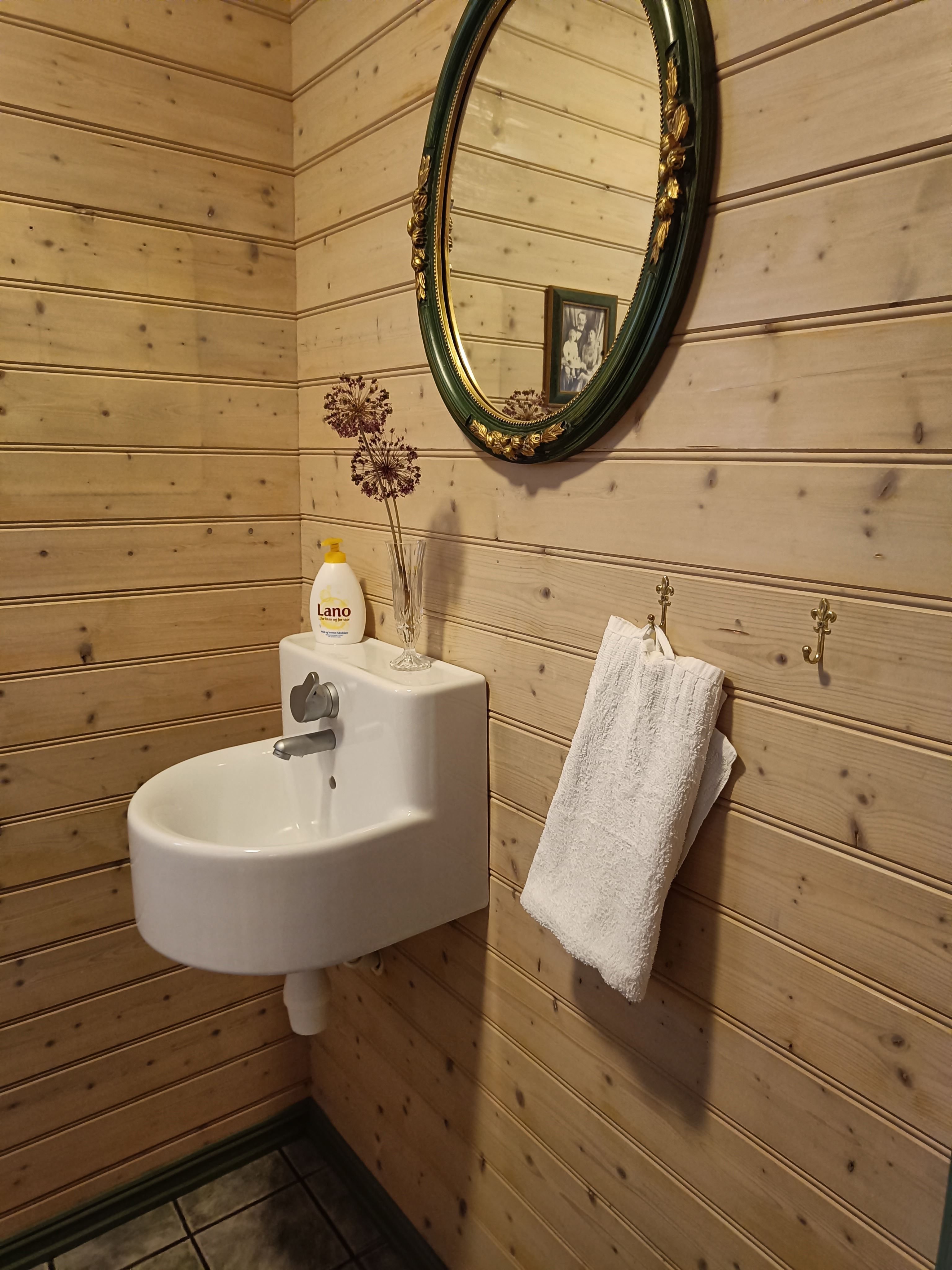 Orheimstunet in Nordfjord - Small bathroom corner with wood-paneled walls, a wall-mounted sink, oval mirror, soap bottle, and towel..