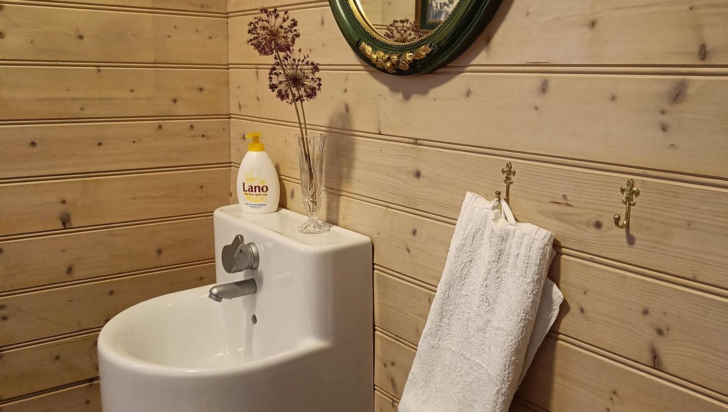 Orheimstunet in Nordfjord - Small bathroom corner with wood-paneled walls, a wall-mounted sink, oval mirror, soap bottle, and towel..