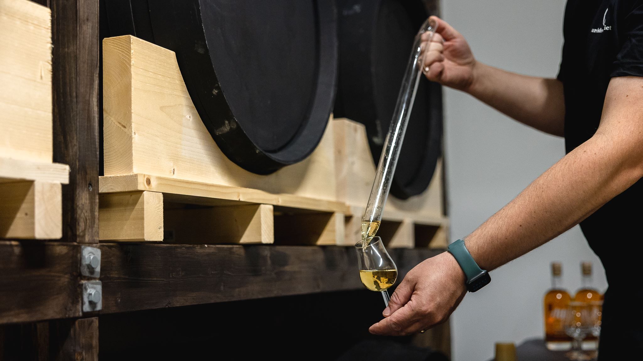 Person draws golden liquor from a barrel with a glass thief and pours it into a tasting glass on a wooden rack.