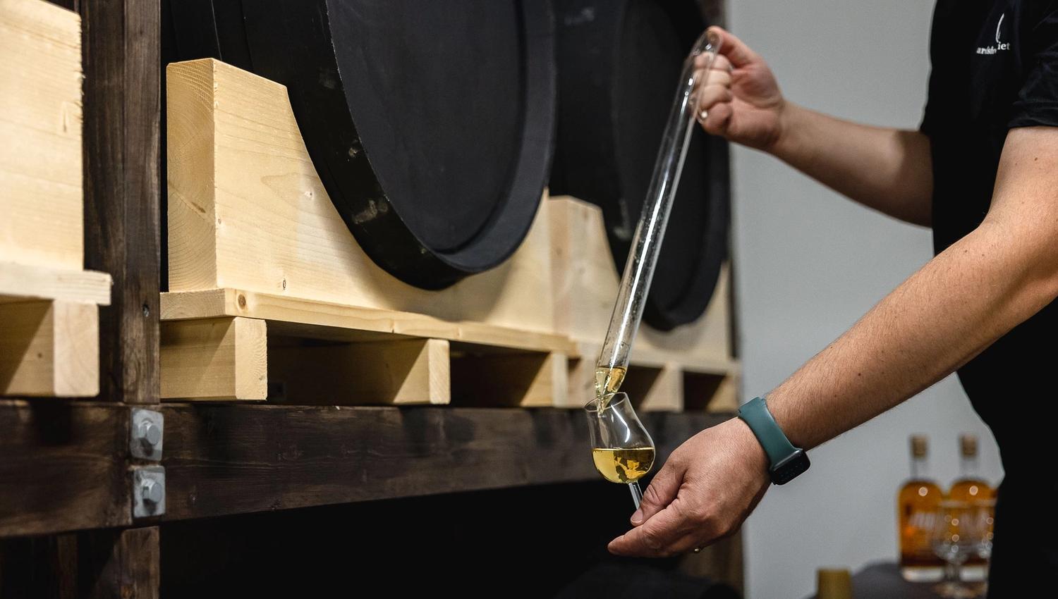 Person draws golden liquor from a barrel with a glass thief and pours it into a tasting glass on a wooden rack.