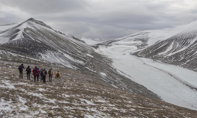 En guide og en gruppe med gjester som går ned en fjellside i forgrunnen med lett snødekte fjell og en isbre i bakgrunnen
