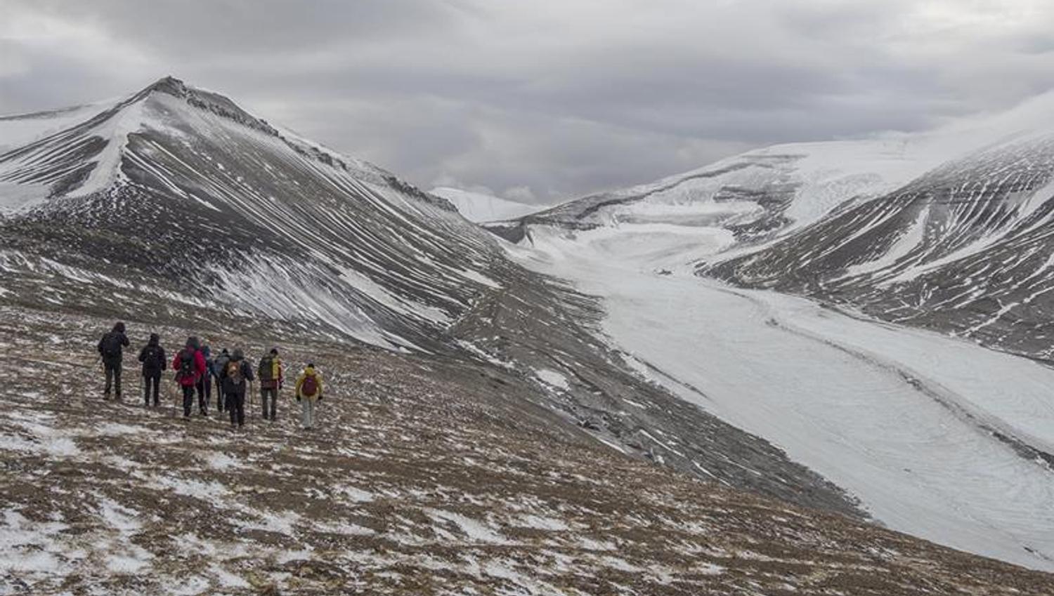En guide og en gruppe med gjester som går ned en fjellside i forgrunnen med lett snødekte fjell og en isbre i bakgrunnen